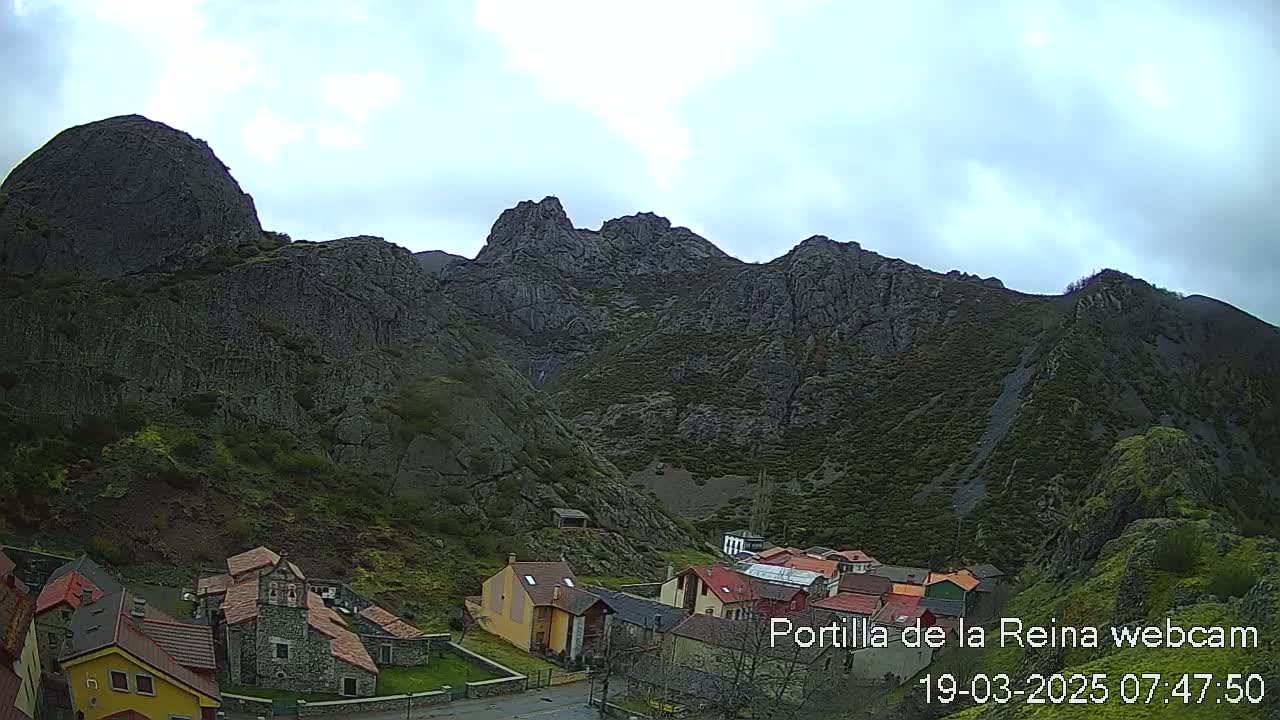 A small village nestled at the base of a large, rocky mountain range under a cloudy sky.