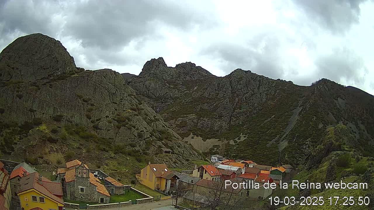 A small village of stone and stucco buildings nestles at the base of a large, rocky mountain range under a cloudy sky.