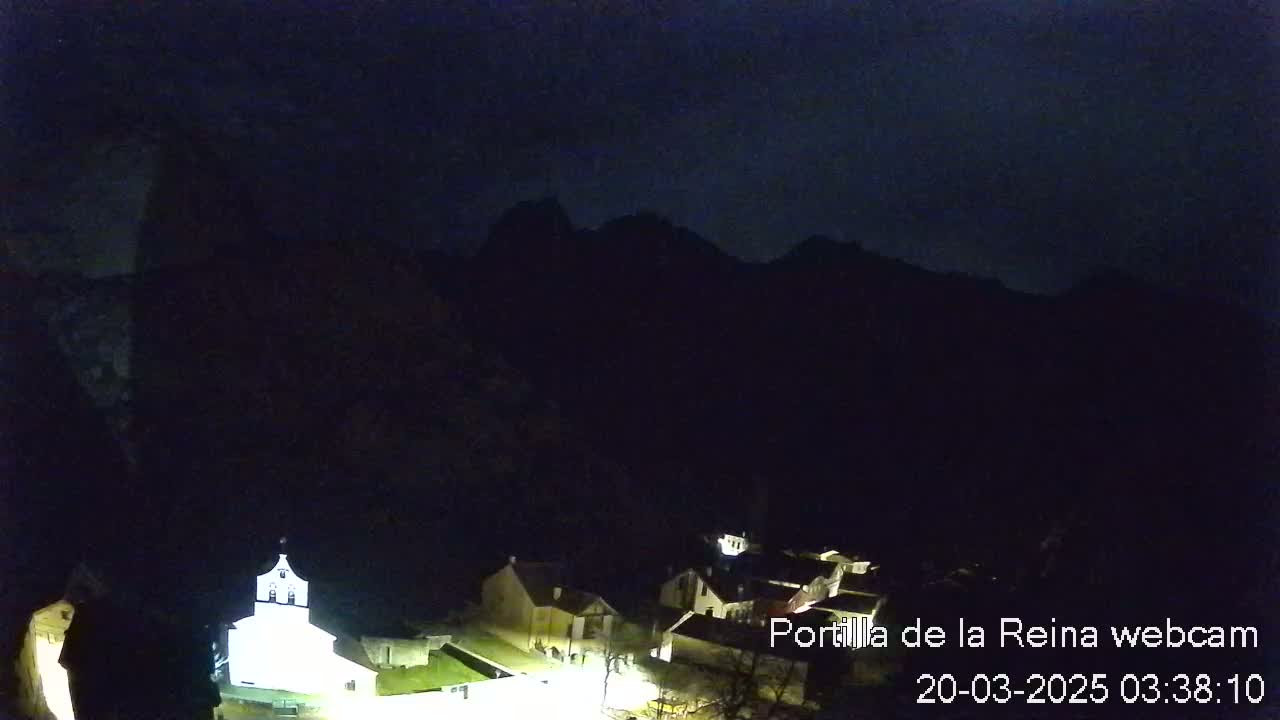 A nighttime view of a small village nestled in a valley, illuminated by faint lights, with dark mountains rising in the background under a cloudy sky.