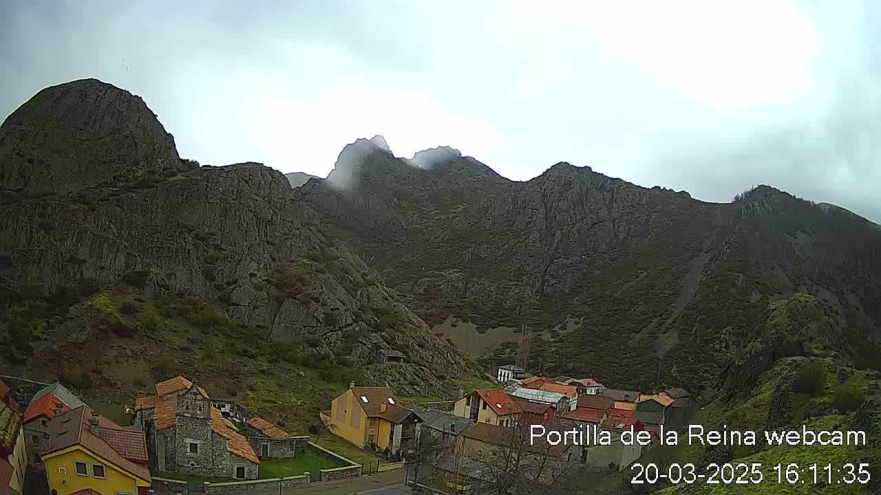 A small village nestled at the base of a large, rocky mountain range under a cloudy sky.