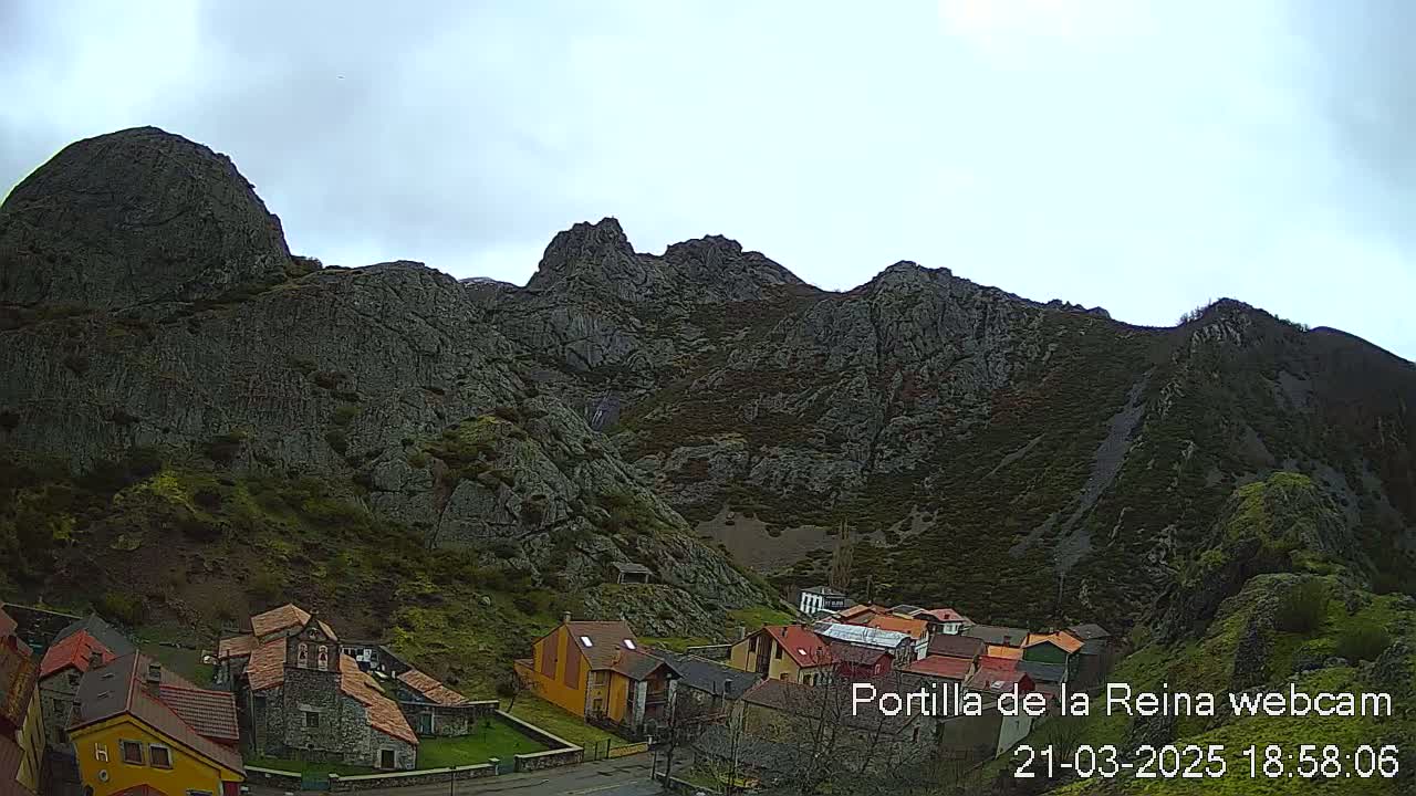 A small village of stone and stucco houses sits at the base of a large, rocky mountain range under an overcast sky.