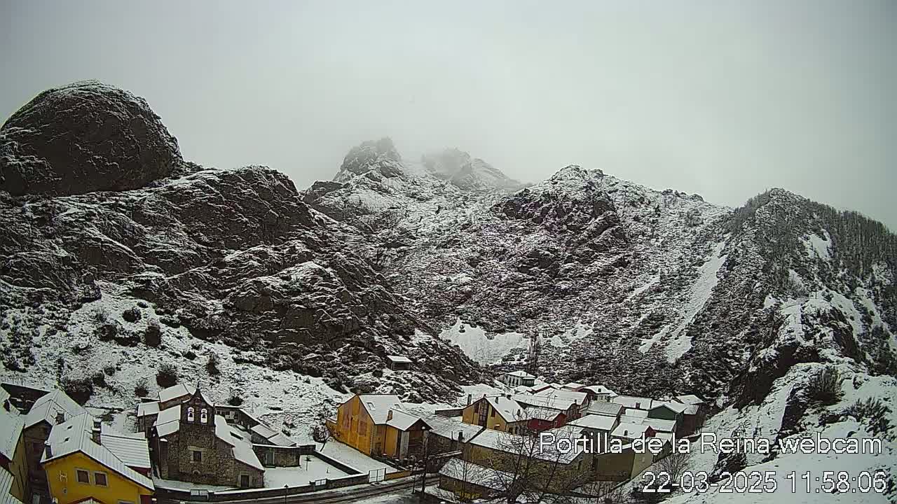 A snow-covered village nestled in a valley between dark, rocky mountains under an overcast sky.