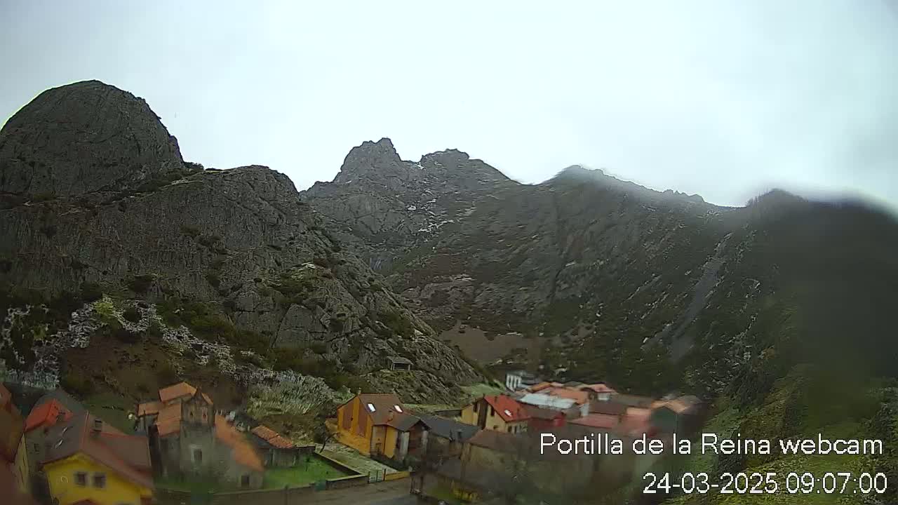 A small village nestled at the base of large, gray, rocky mountains under an overcast sky.