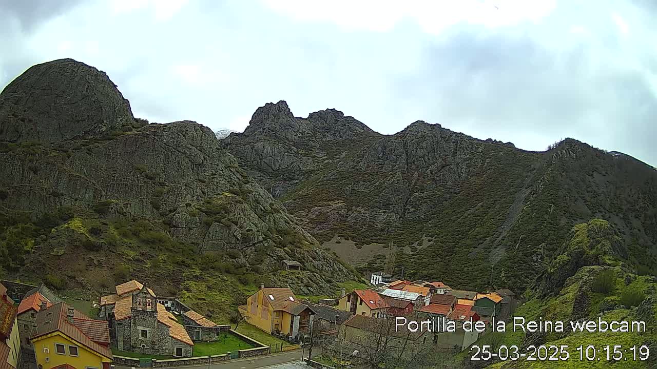 A small village nestled at the base of a large, rocky mountain range under a cloudy sky.