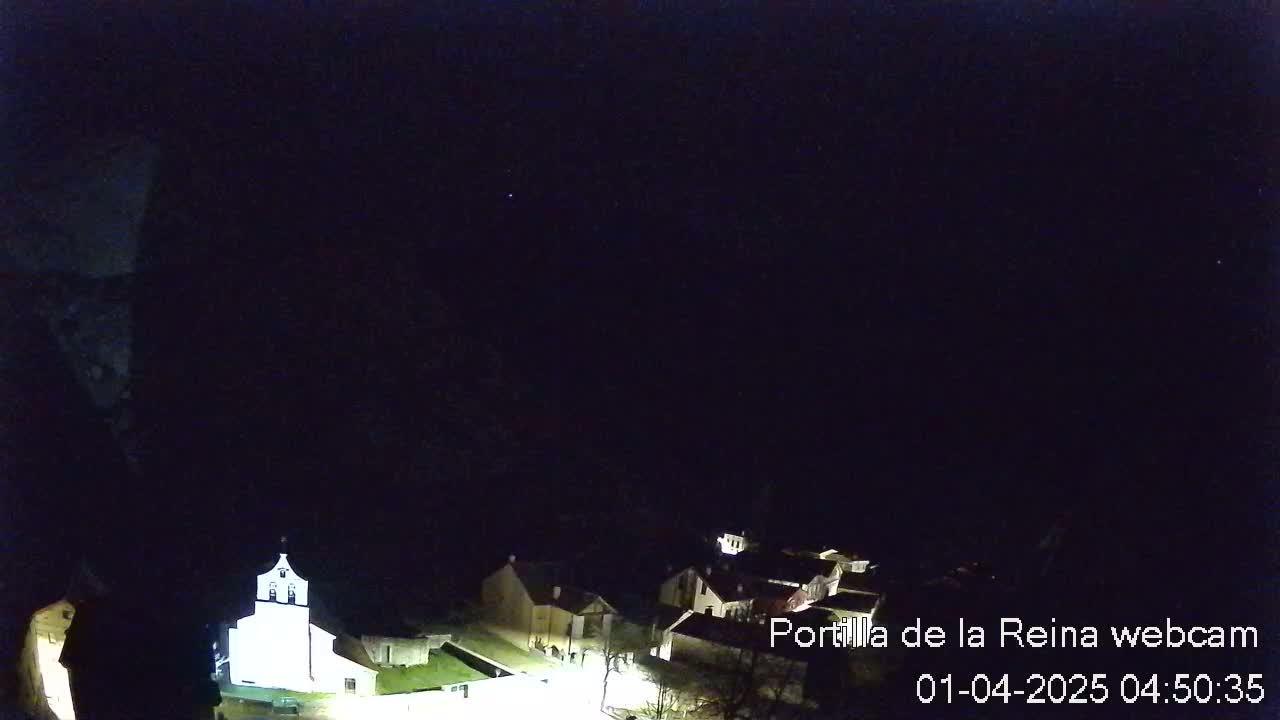 A small, dimly lit village is nestled at the base of a dark mountain under a clear, starlit night sky.
