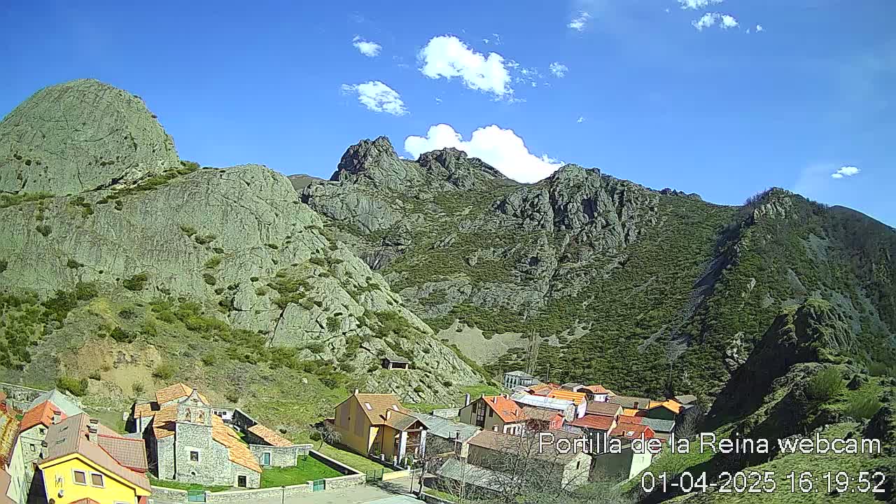 A small village nestled in a valley between rocky, green-covered mountains under a mostly sunny sky with a few fluffy clouds.