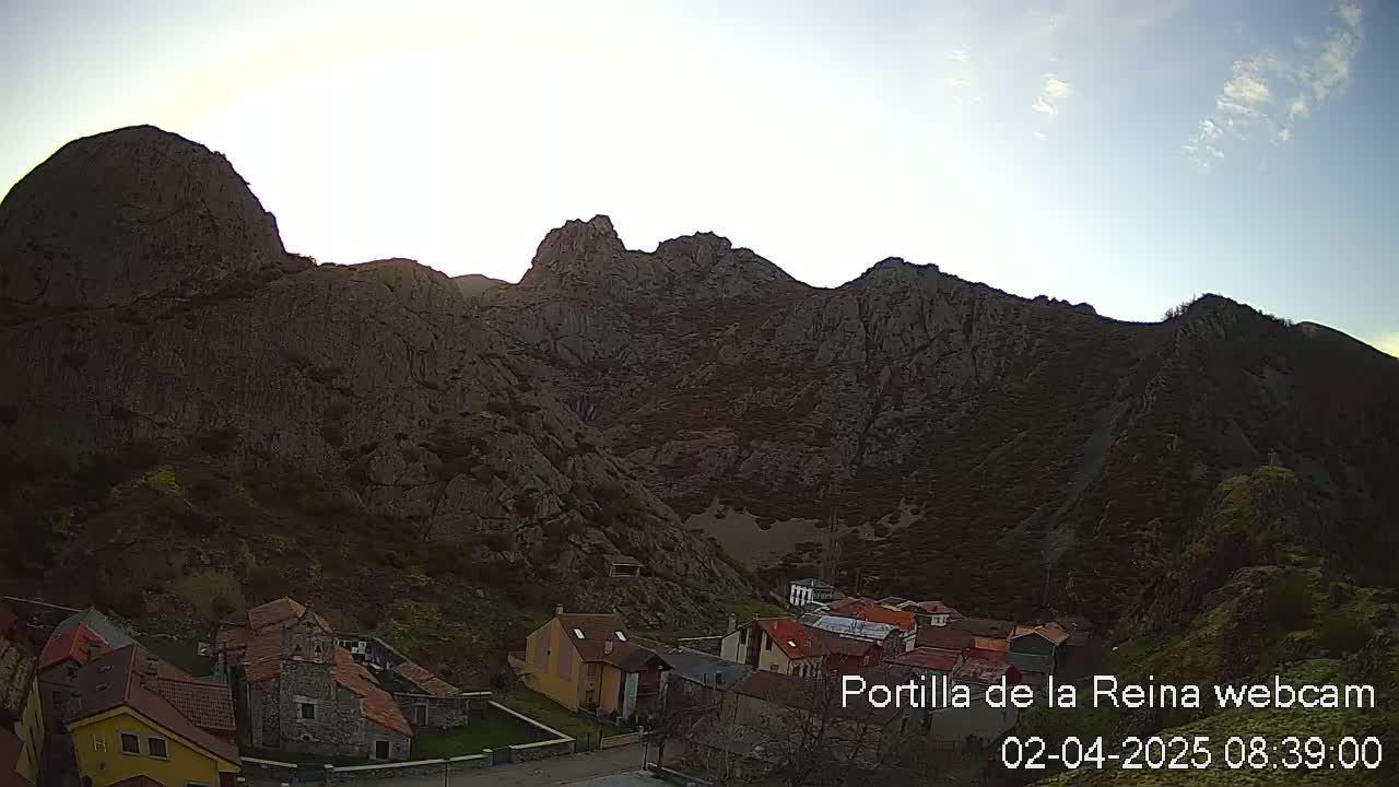 A small village nestled at the base of a large, rocky mountain range under a mostly clear, bright sky.