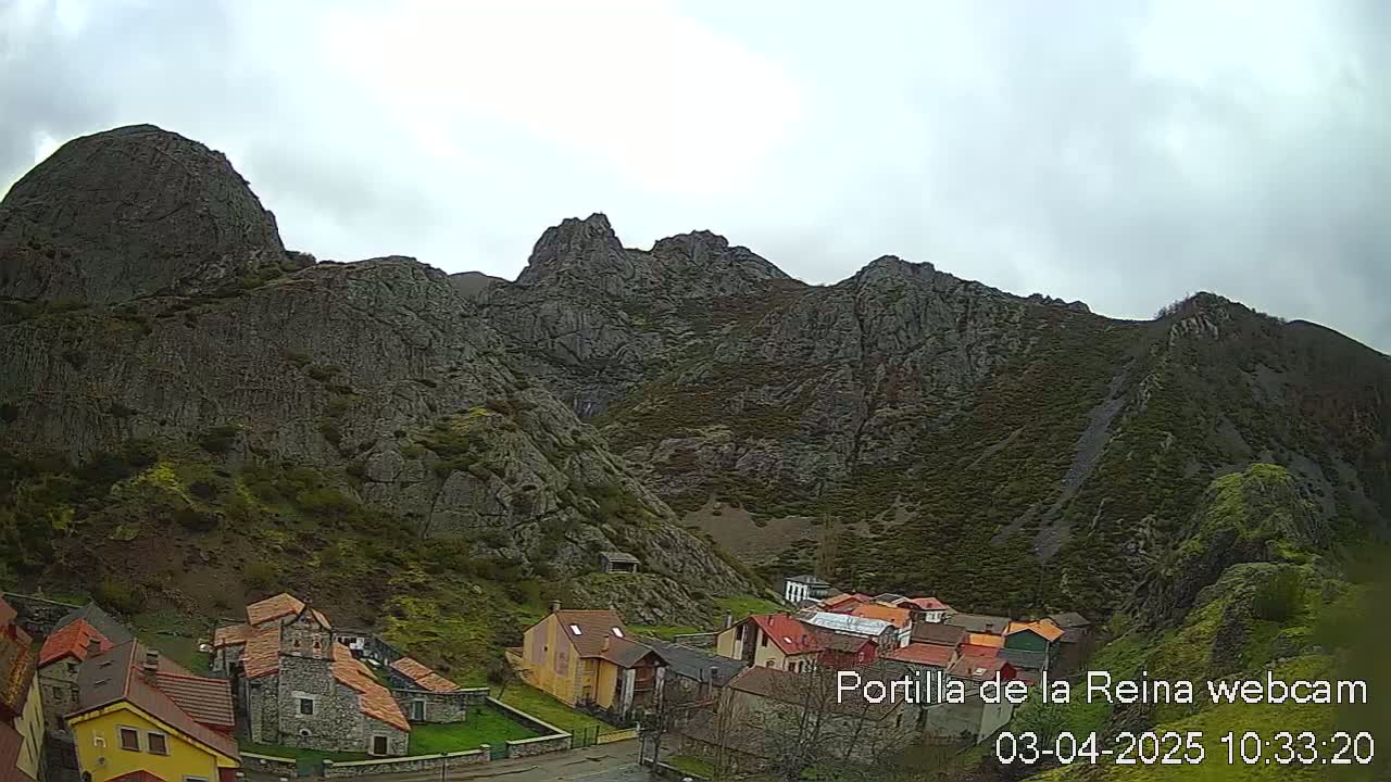 A small village nestled at the base of a large, rocky mountain range under an overcast sky.
