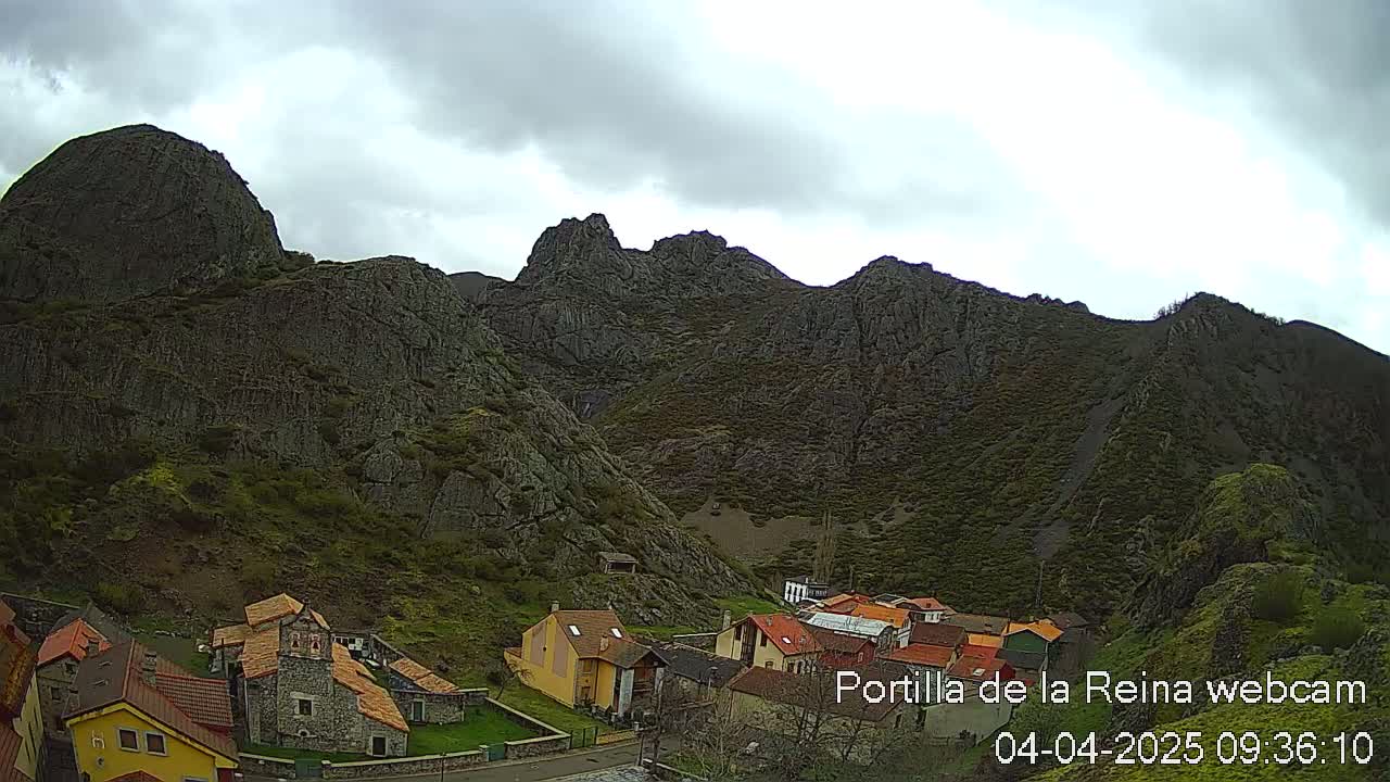 A small village nestled at the base of a large, rocky mountain range under a cloudy sky.