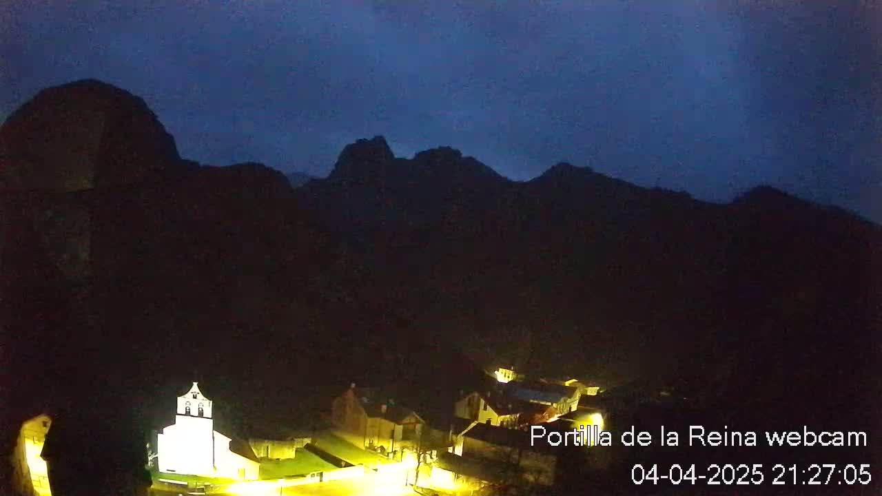 A small, illuminated village nestled in a valley at night, surrounded by dark mountains under a twilight sky.