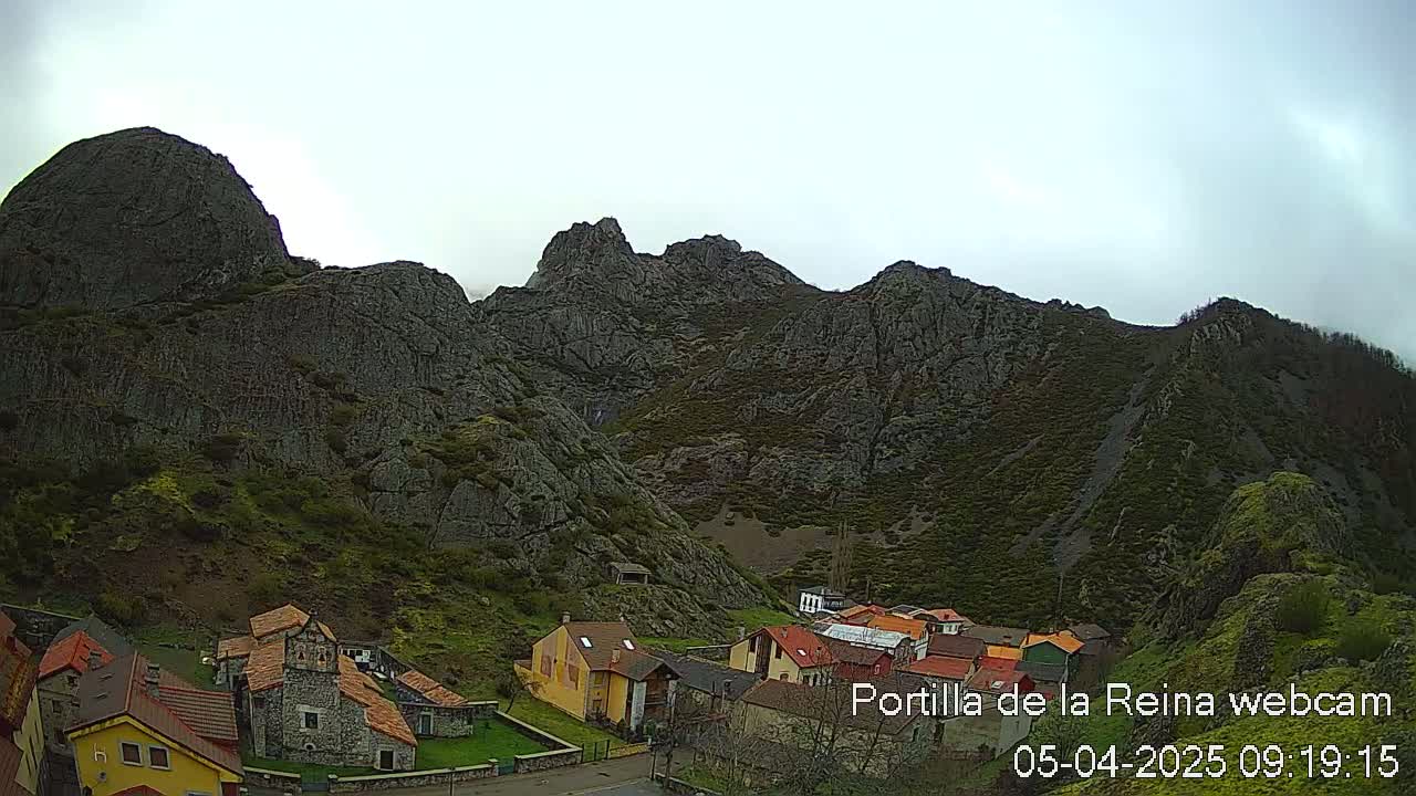 A small village nestled at the base of a large, rocky mountain range under an overcast sky.