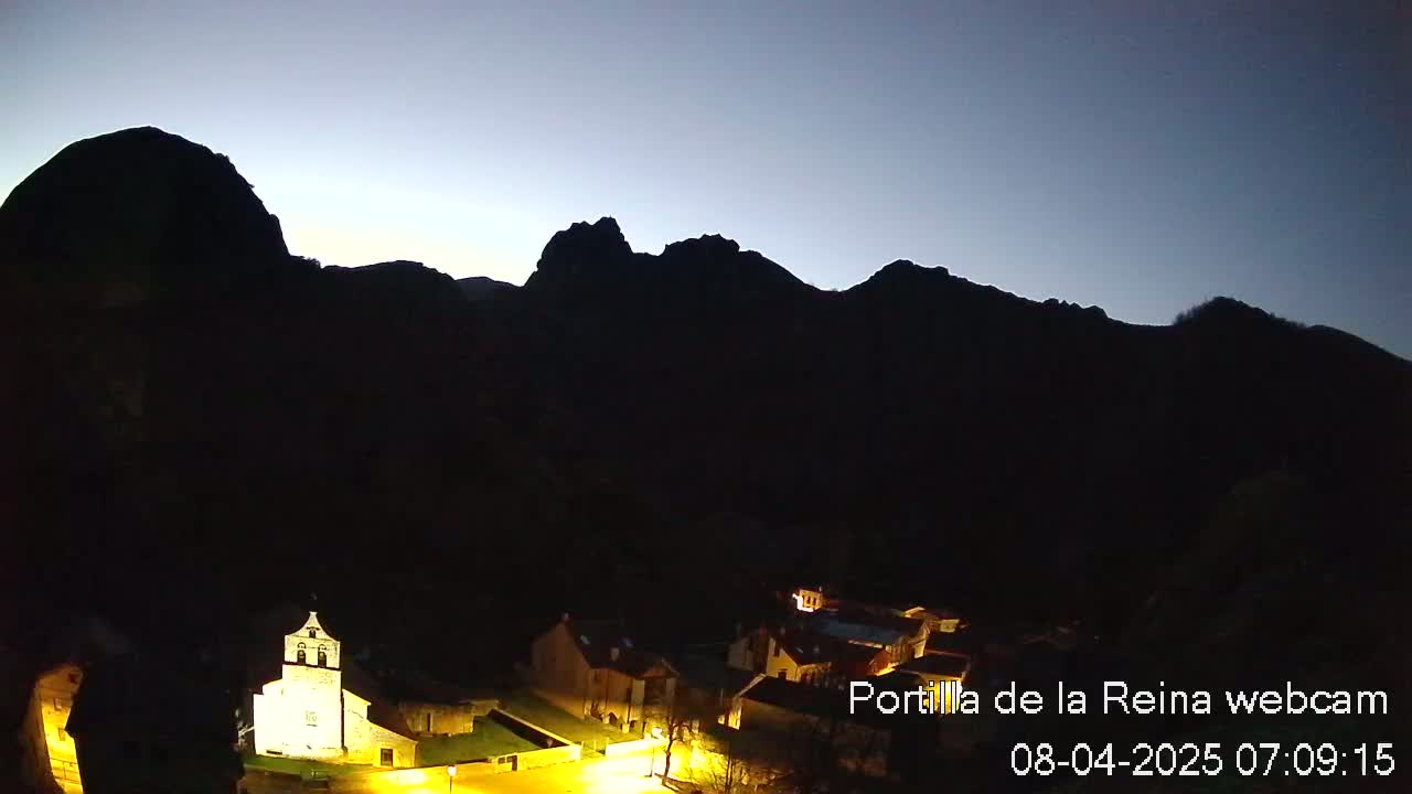 A small, illuminated village nestled at the base of dark, silhouetted mountains under a twilight sky.
