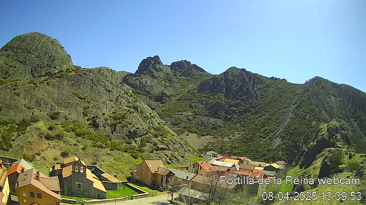 A small village nestled in a valley surrounded by rocky, green-covered mountains under a clear blue sky.