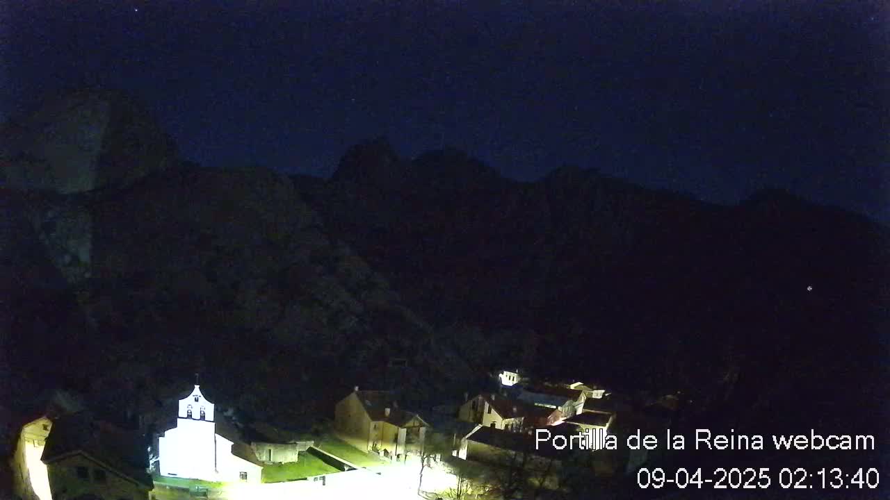 A small, illuminated village nestled in a dark valley under a clear, starlit night sky, surrounded by mountains.