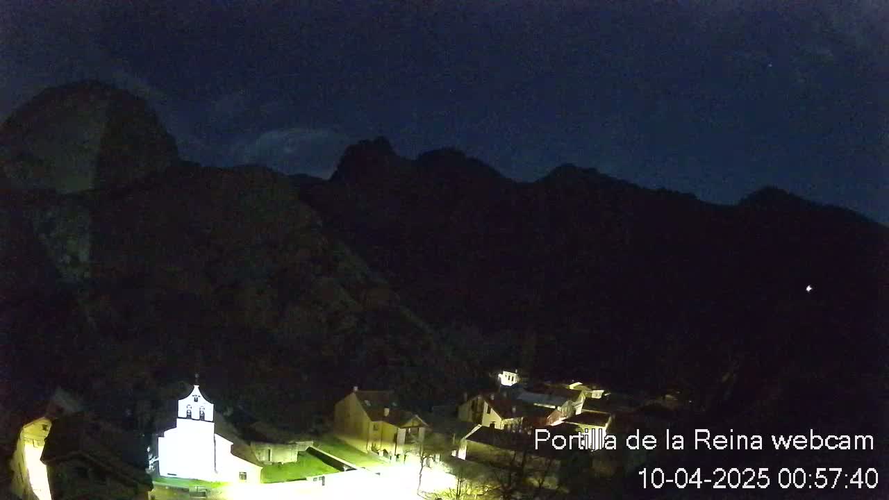 A small, illuminated village nestled at the base of dark mountains under a clear, night sky.