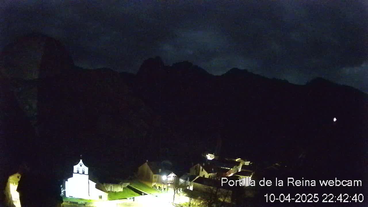 A small, illuminated village nestled at the base of dark mountains under a nighttime sky with some clouds.