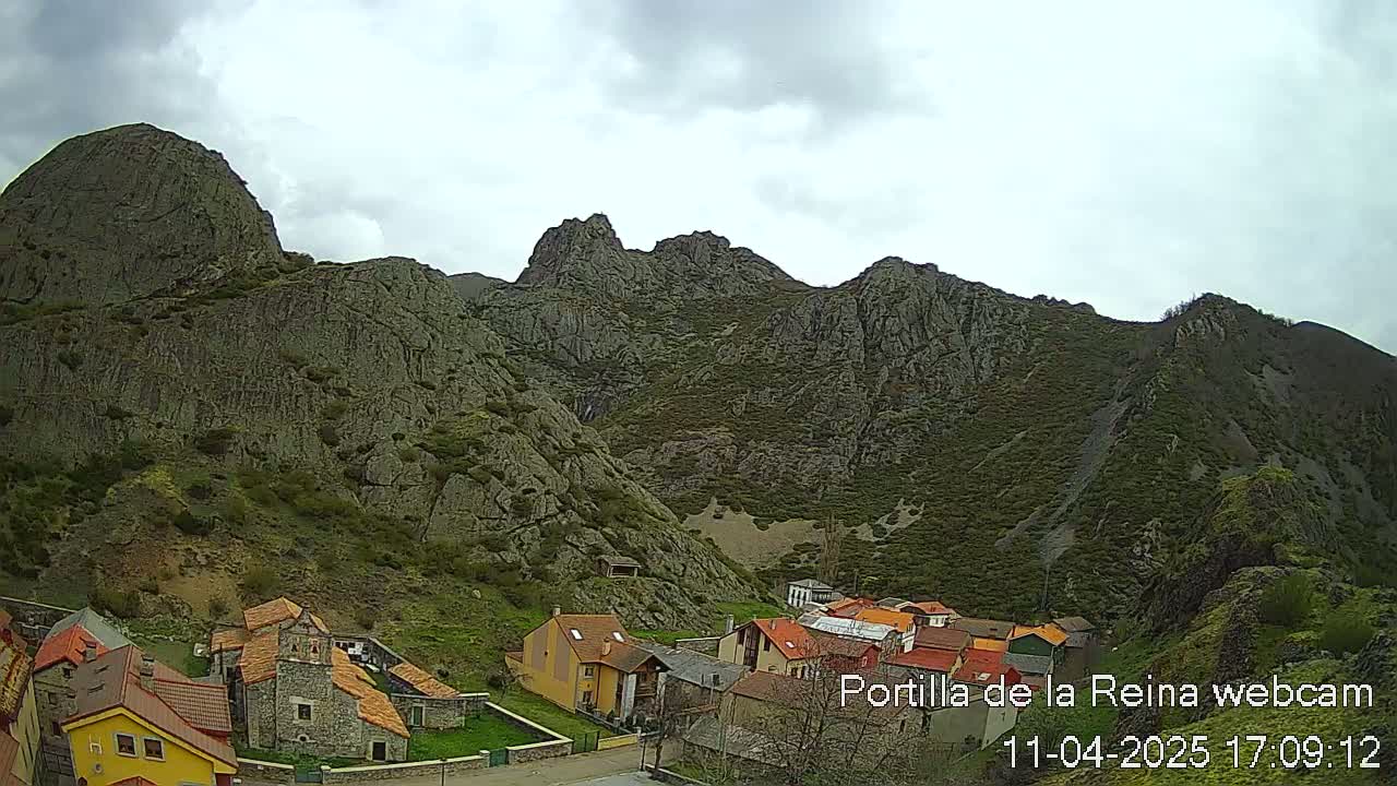 A small village nestled at the base of a large, rocky mountain range under a cloudy sky.