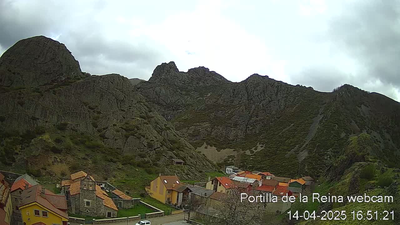 A small village nestled at the base of imposing, dark gray, rocky mountains under a cloudy sky.