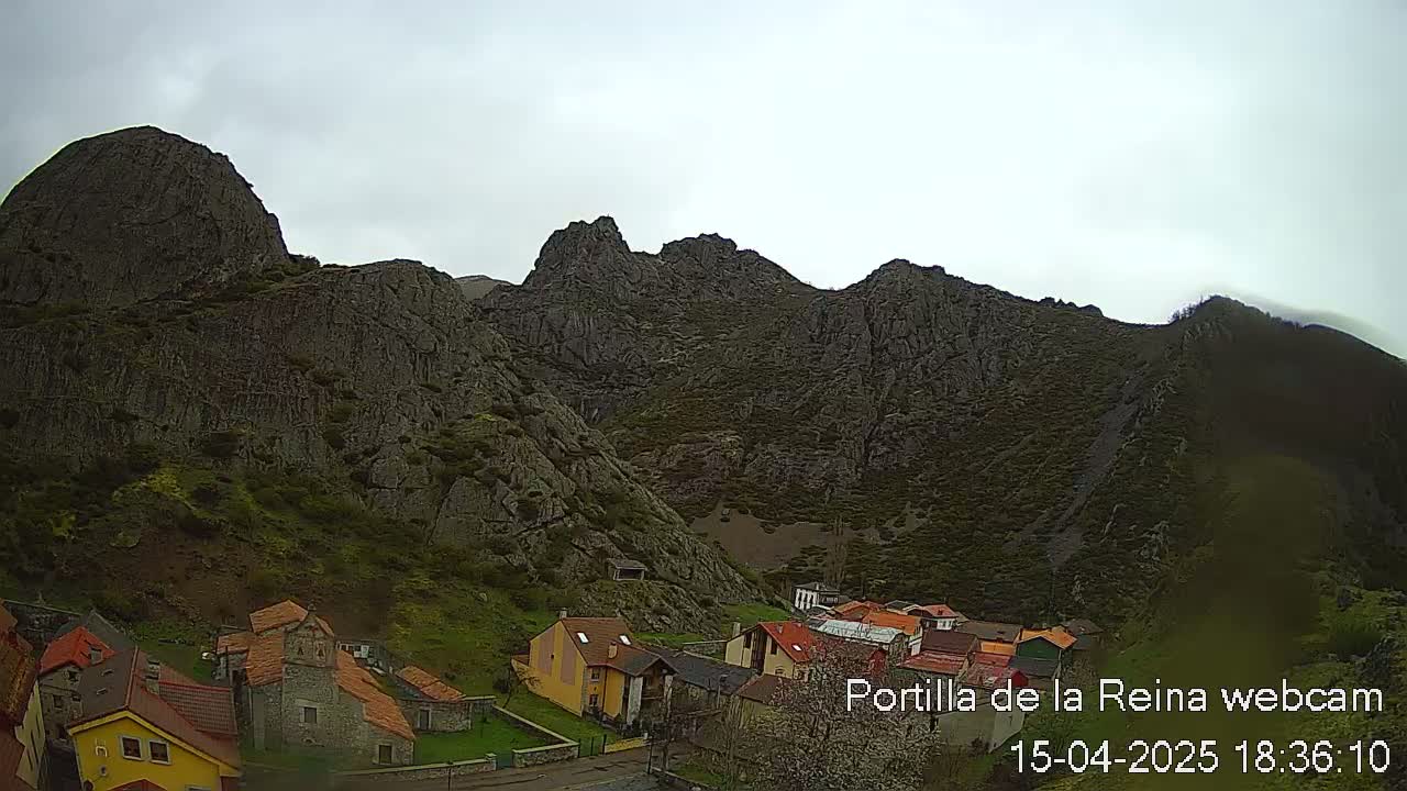 A small village nestled at the base of a large, rocky mountain range under an overcast sky.