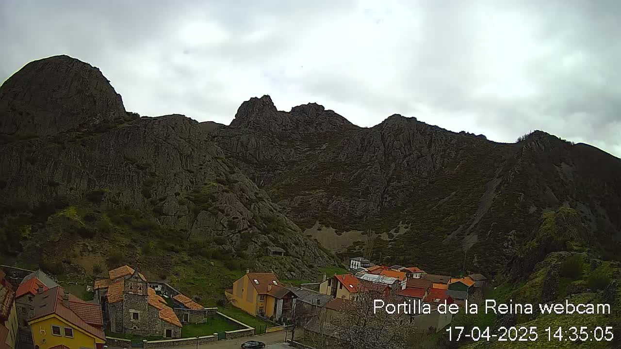 A small village nestled at the base of a large, dark, rocky mountain range under a cloudy sky.
