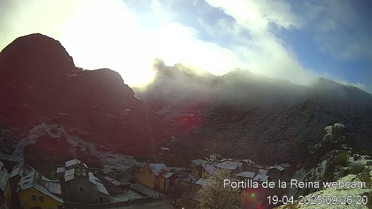 A small village nestled in a valley between snow-dusted mountains under a partly sunny sky with some fog.