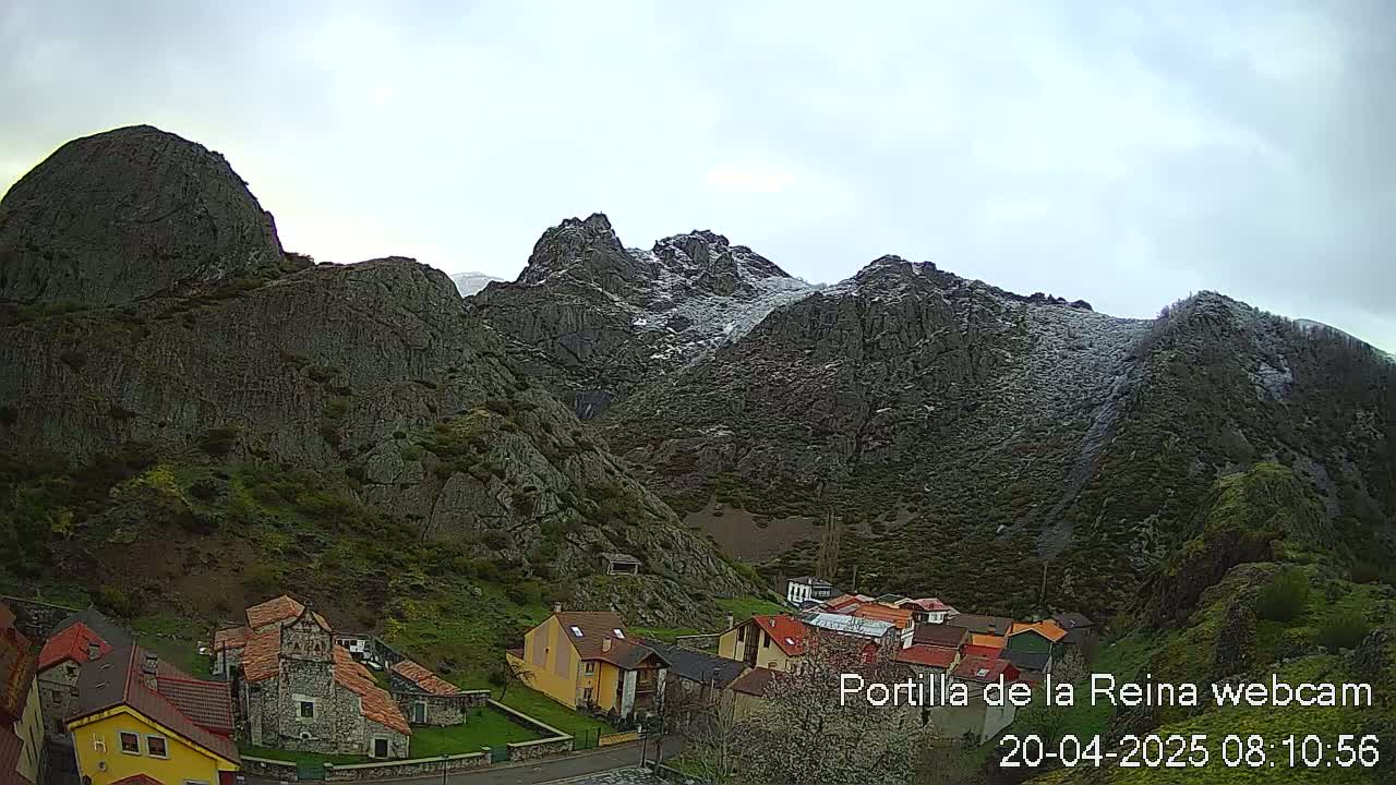 A small village nestled in a valley between snow-dusted, rocky mountains under an overcast sky.