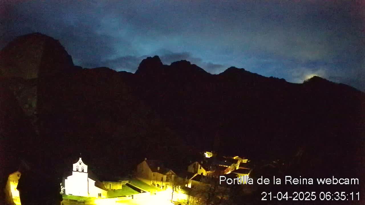 A small village with illuminated buildings is nestled at the base of dark, silhouetted mountains under a twilight sky.
