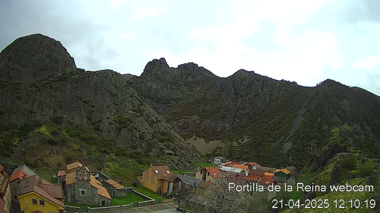 A small village nestled at the base of a large, rocky mountain range under a mostly cloudy sky.