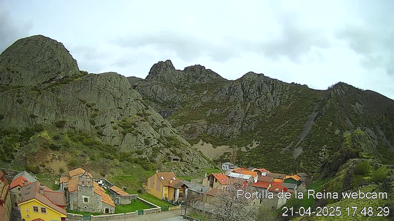 A small village nestled at the base of a large, rocky mountain range under a cloudy sky.