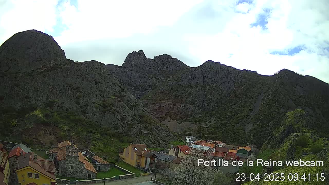 A small village nestled at the base of a large, dark, rocky mountain range under a partly cloudy sky.