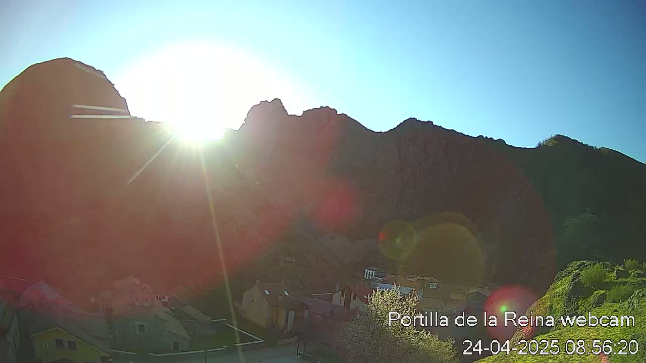 A bright sun shines over a village nestled at the base of a mountain range on a clear day.