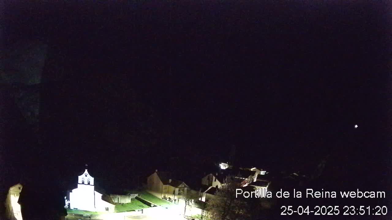 A nighttime, long shot of a small village with a brightly lit church, under a clear, dark sky.