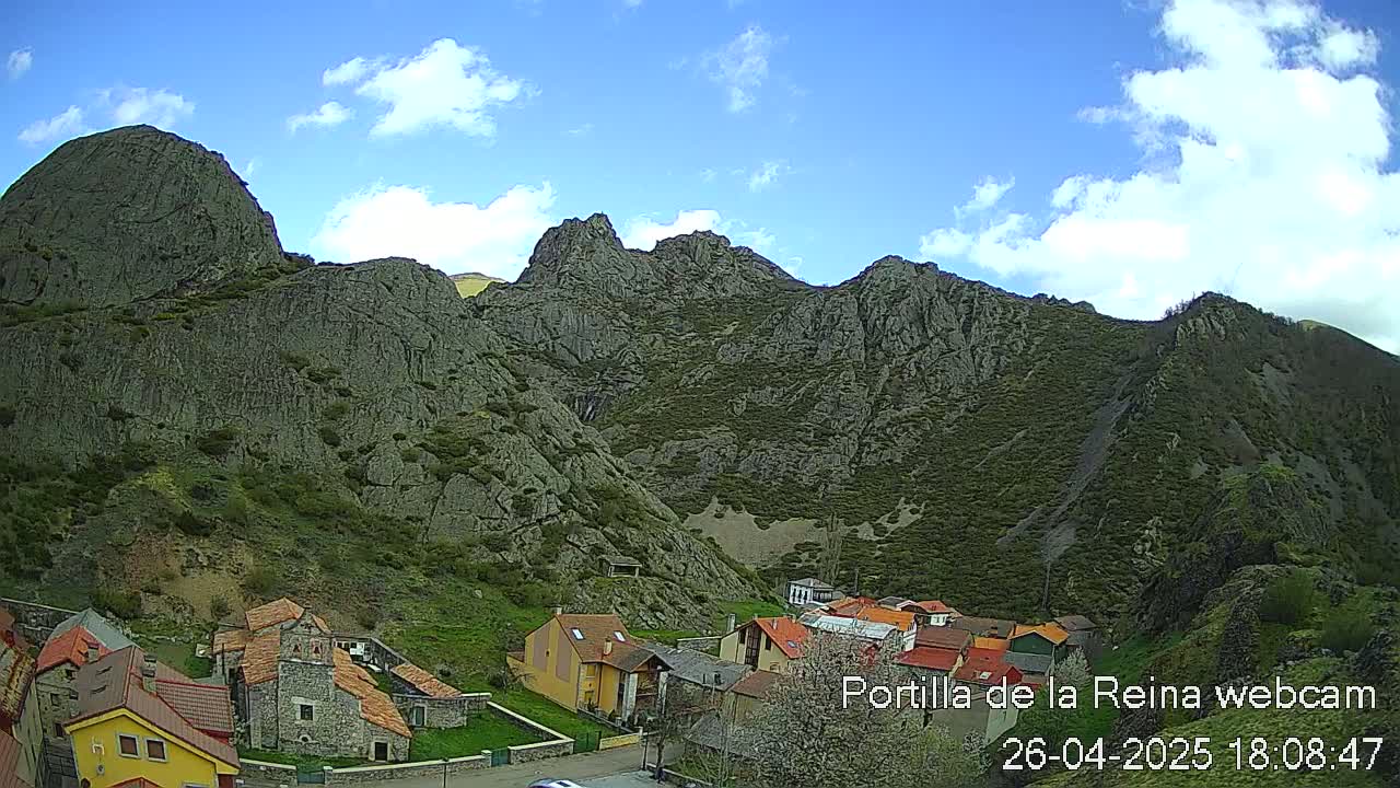 A small village nestled at the base of a large, rocky mountain range under a partly cloudy blue sky.
