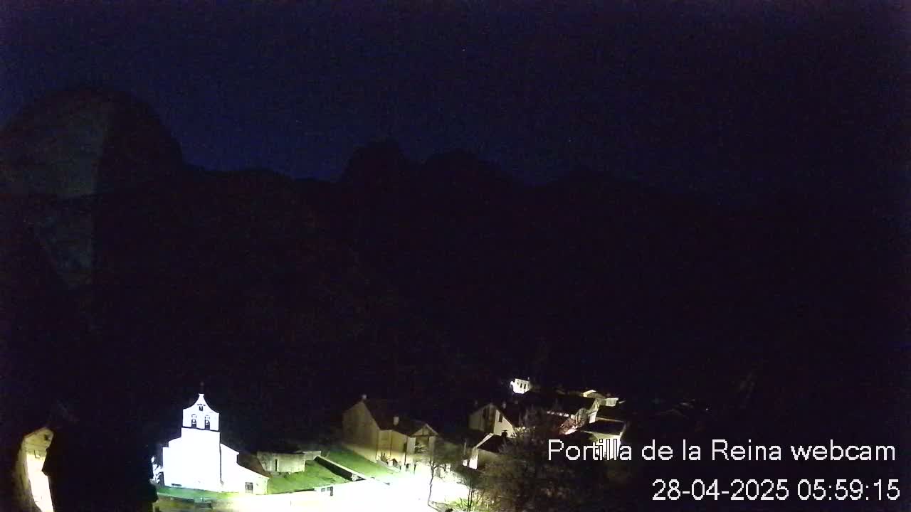 A small, dark village nestled in a valley under a night sky is illuminated by a few lights, with a church prominently lit in the foreground.