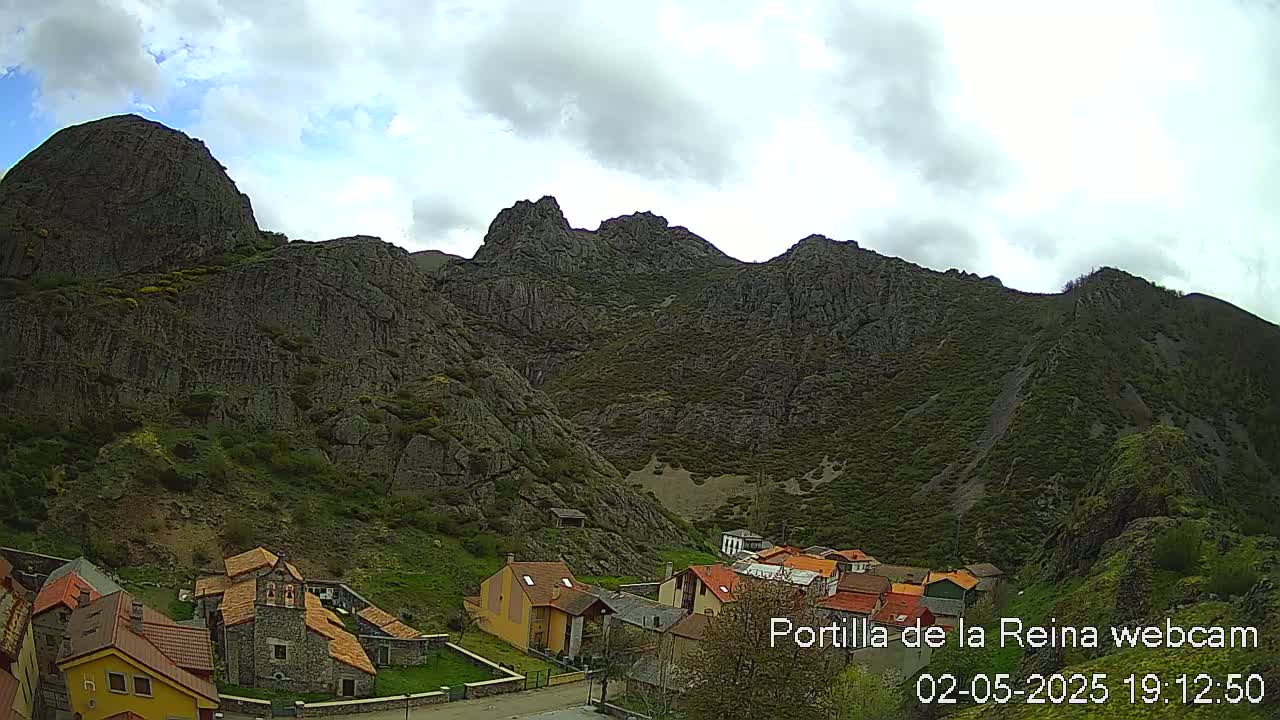 A small village nestled at the base of a large, rocky mountain range under a partly cloudy sky.