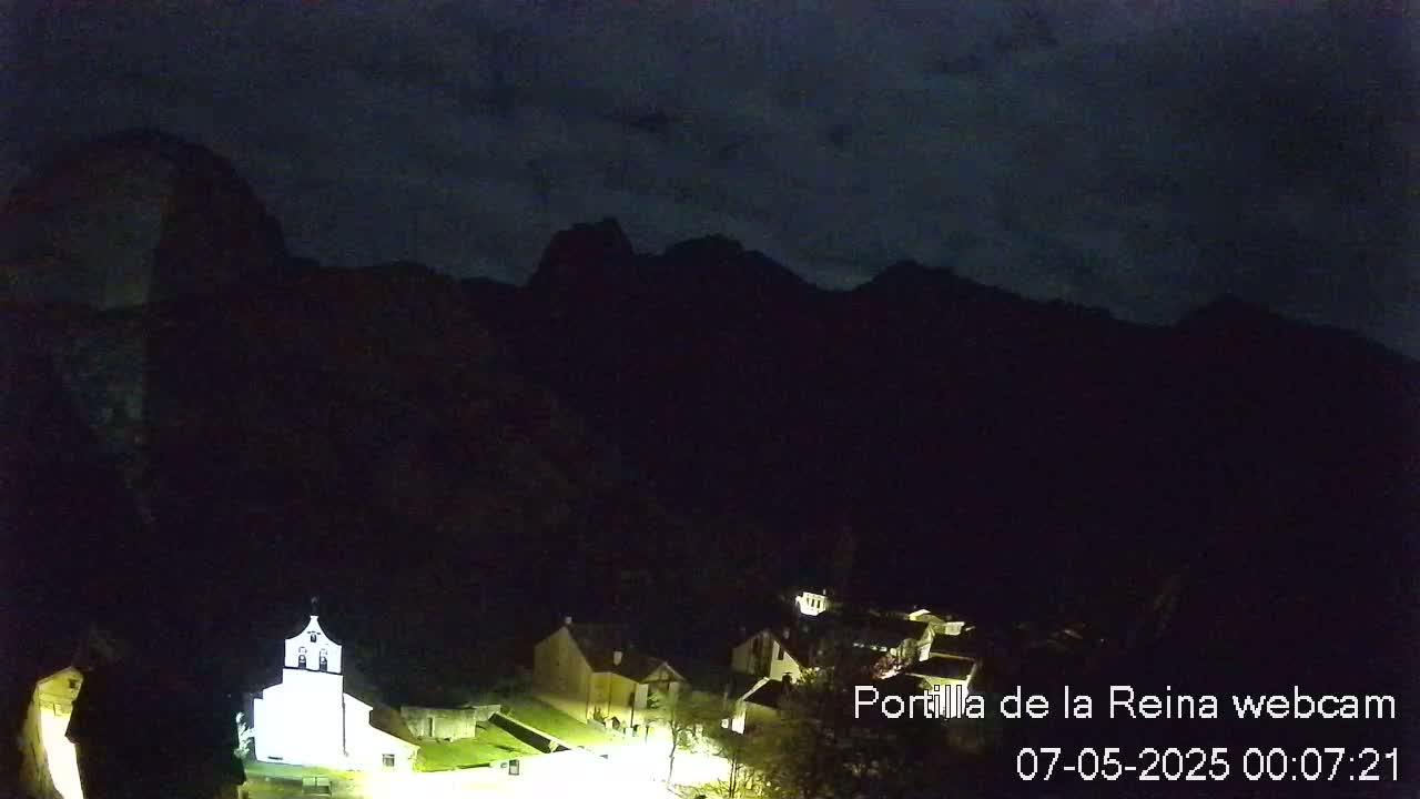 A small, dark village with dimly lit buildings is nestled at the base of silhouetted mountains under a dark, cloudy night sky.