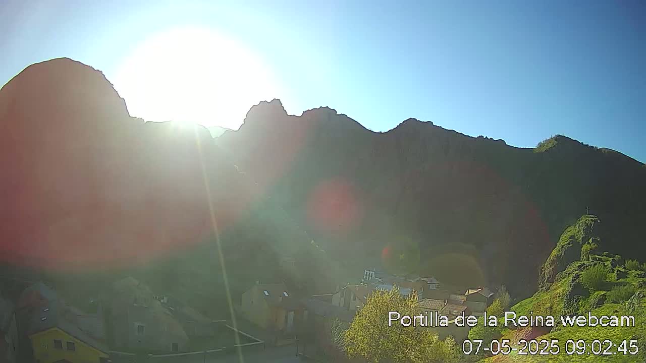 A bright sun shines over a village nestled at the base of a large, dark mountain range on a clear day.