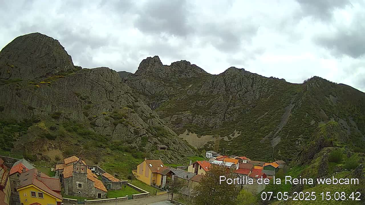 A small village nestled at the base of a large, rocky mountain range under a cloudy sky.
