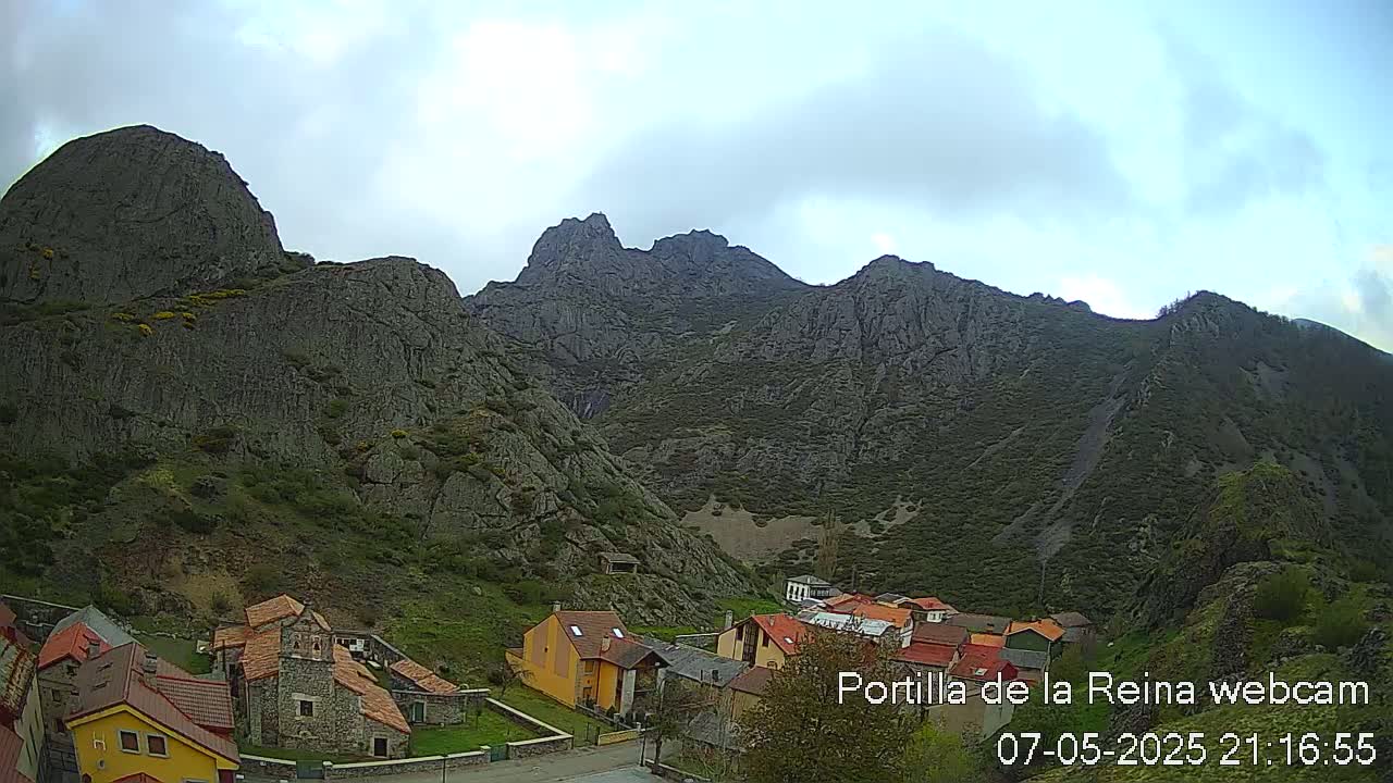A small village of stone and stucco houses nestles at the base of a large, gray, rocky mountain range under a cloudy sky.