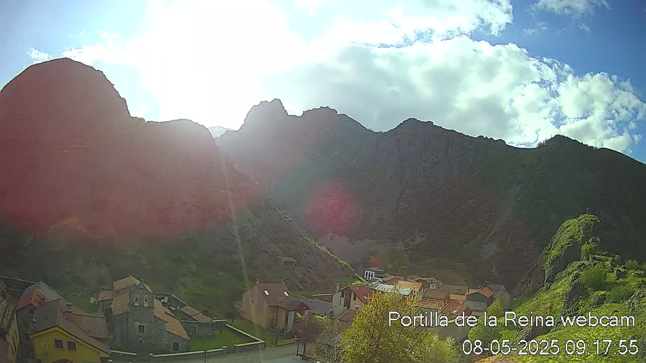 A sunny day reveals a village nestled at the base of large, dark mountains under a partly cloudy sky.