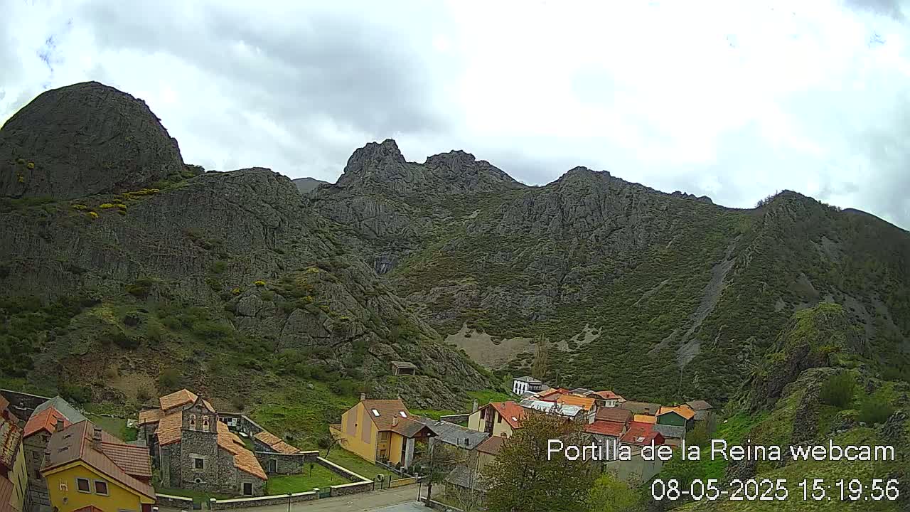 A small village nestled at the base of a large, rocky mountain range under a cloudy sky.