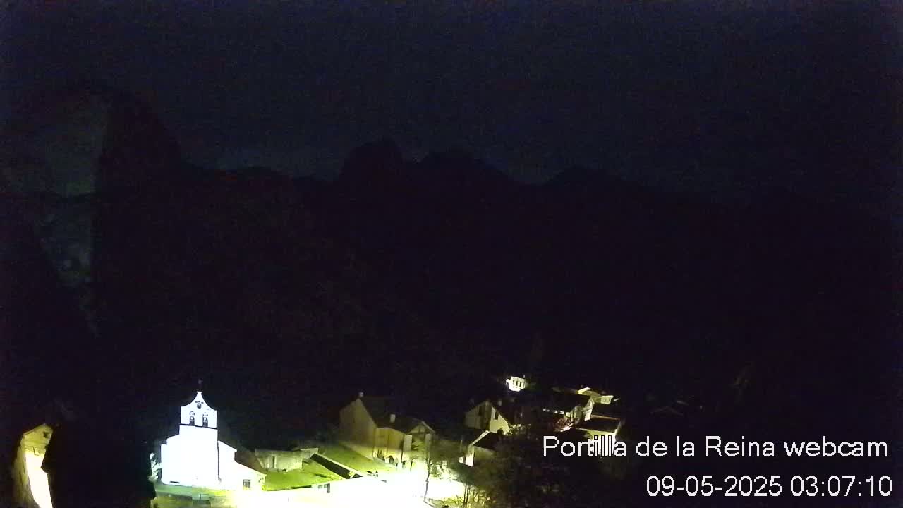 A small, dimly lit village nestled at the base of dark mountains under a nighttime sky.
