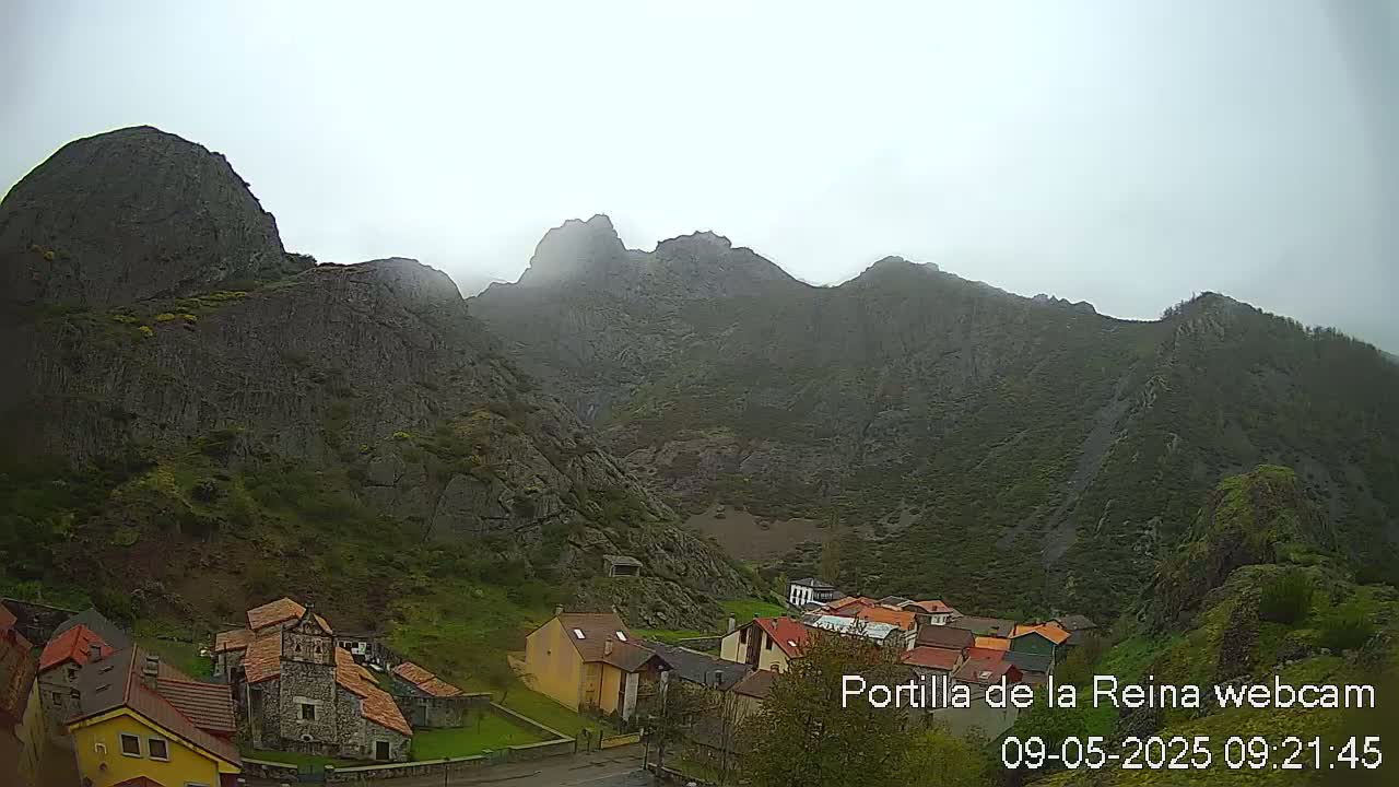 A small village nestled at the base of a large, mist-shrouded mountain range on an overcast day.