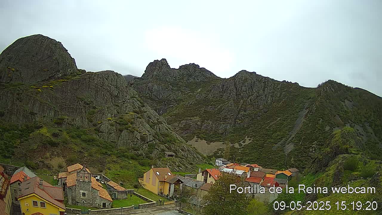 A small village nestled in a valley surrounded by rocky, green mountains under an overcast sky.