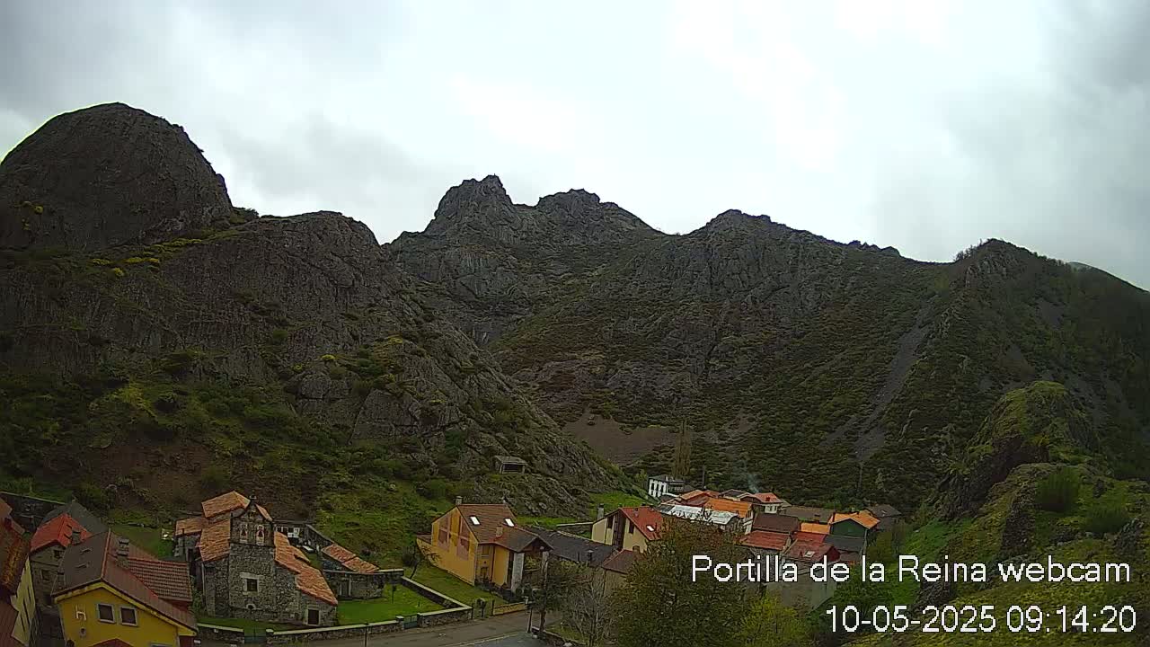A small village nestled at the base of a large, rocky mountain range under an overcast sky.