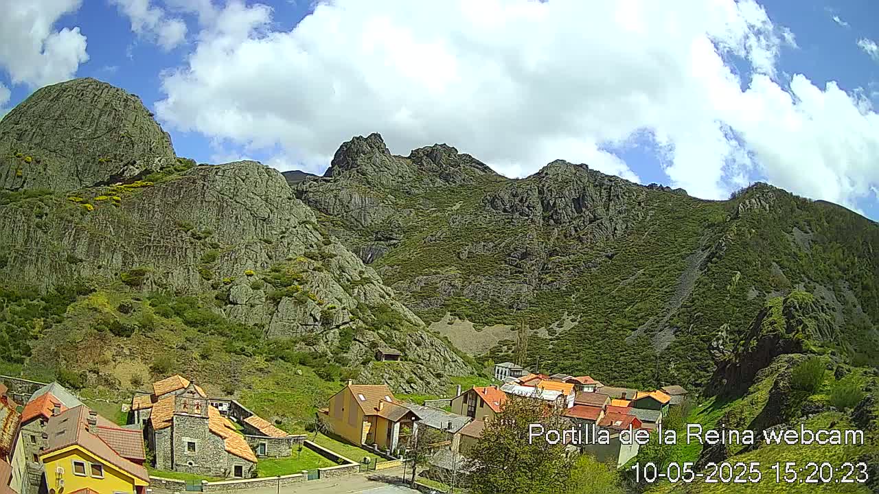A small village nestled at the base of a large, rocky mountain range under a partly cloudy sky.