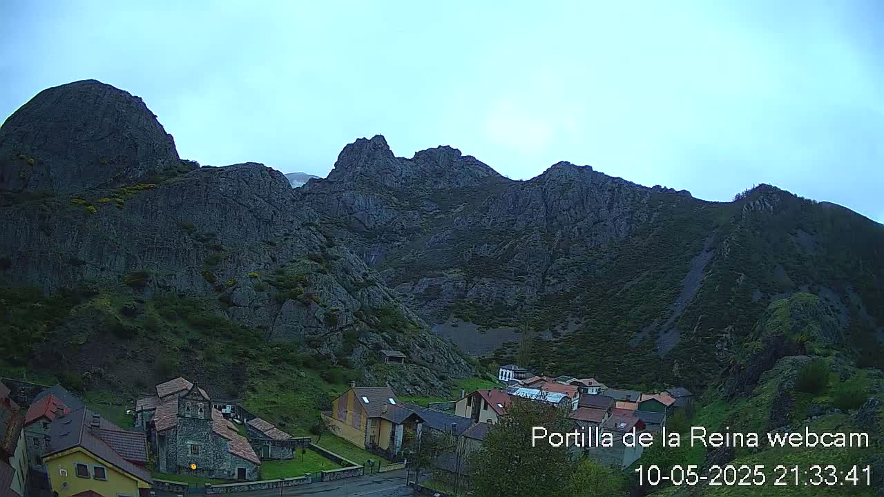 A small village nestled at the base of large, dark, rocky mountains under a clear, light-blue sky.