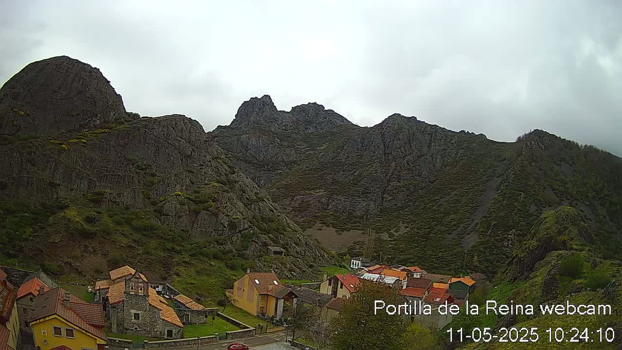A small village of stone and stucco houses nestles at the base of a large, rocky mountain range under an overcast sky.