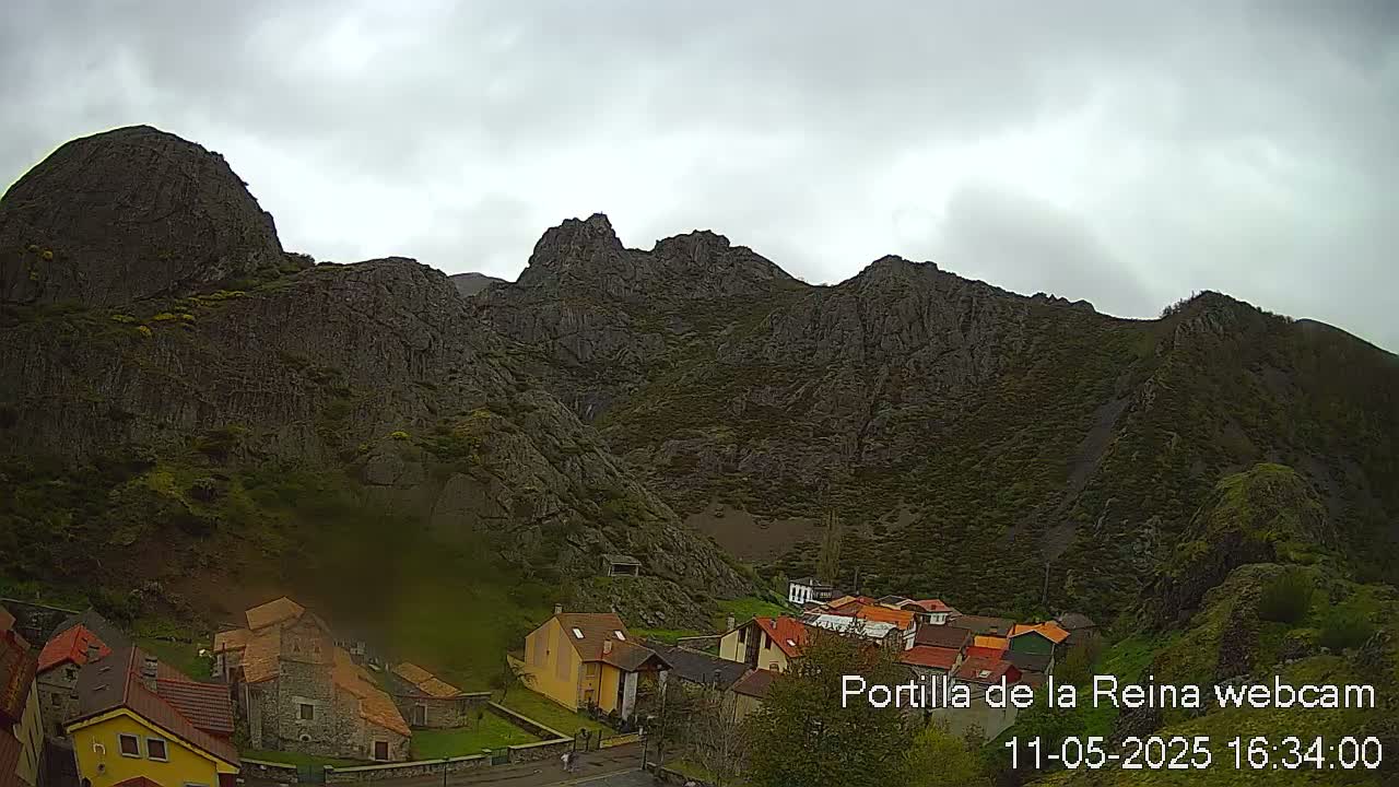 A small village nestled at the base of a large, rocky mountain range under an overcast sky.