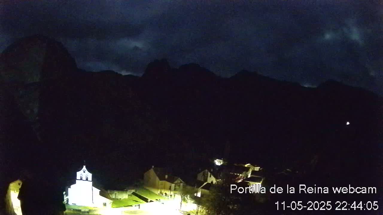 A small village nestled at the base of dark mountains under a cloudy night sky.
